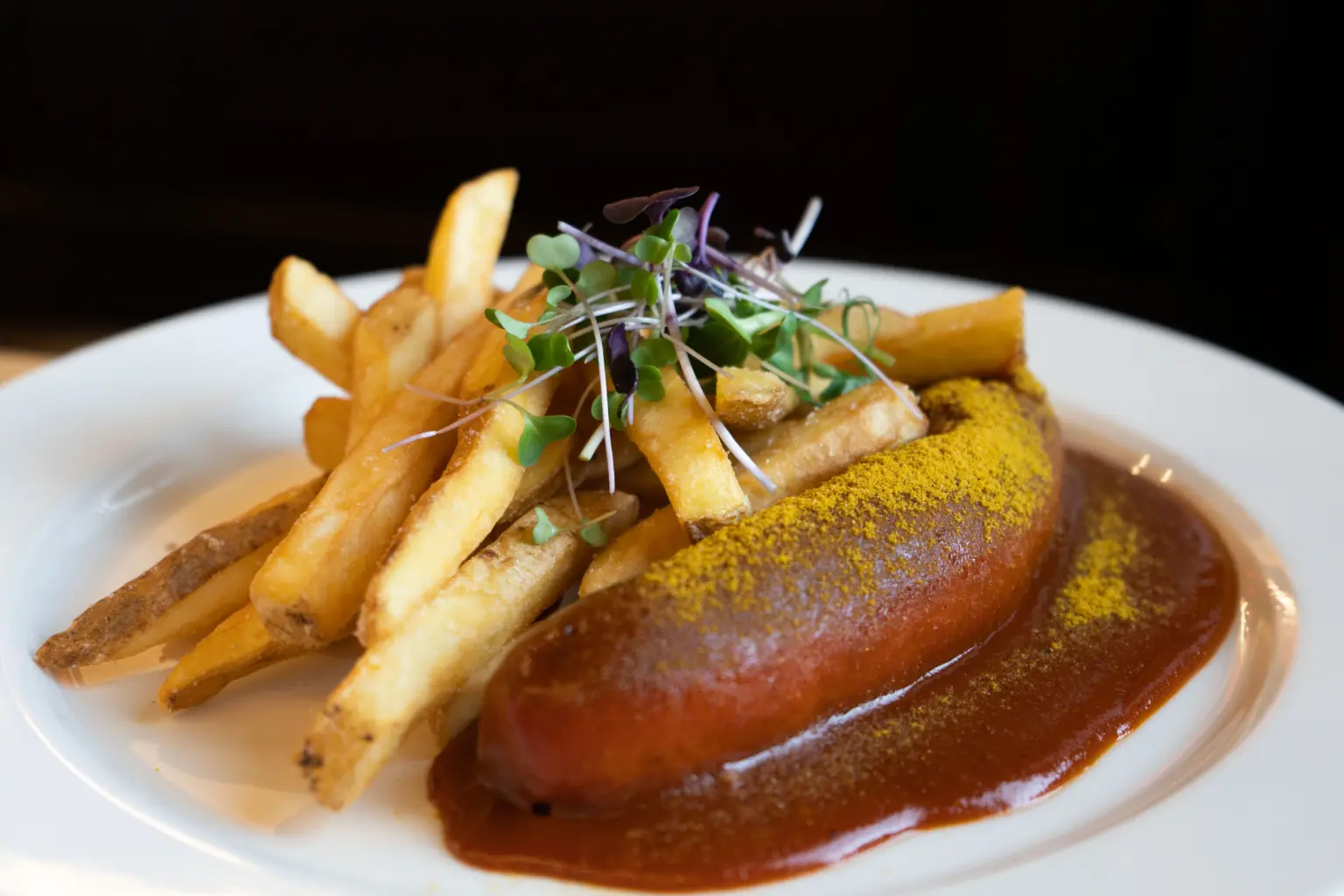 Herzhafte Currywurst in dunkler Sauce, bestäubt mit Currypulver, serviert mit goldgelben Pommes frites auf einem weißen Teller.