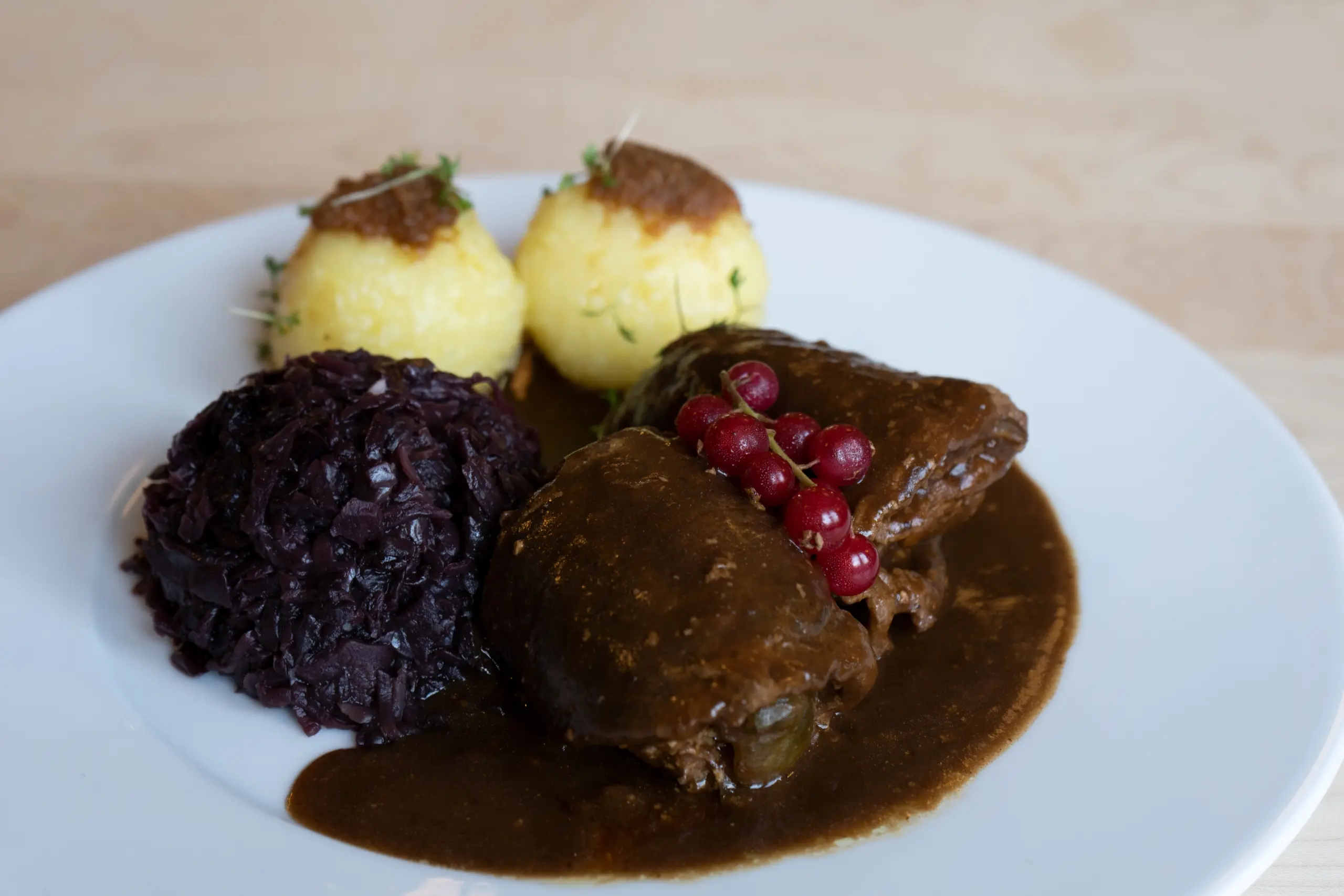 Zwei gefüllte Rinderrouladen in dunkler Bratensauce, serviert mit zwei Kartoffelklößen, Apfelrotkohl und einer Garnitur aus roten Johannisbeeren auf einem weißen Teller.
