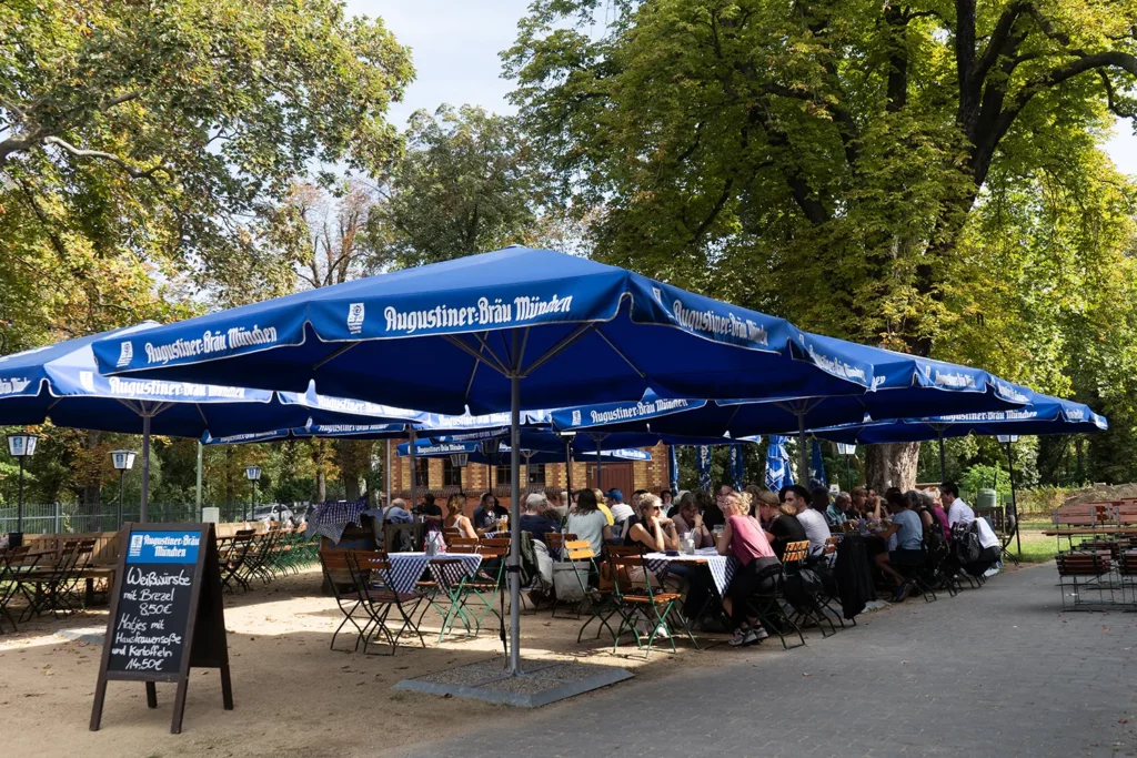 Einladender Biergarten an einem sonnigen Tag mit Gästen unter großen, blauen Augustiner-Bräu Sonnenschirmen.