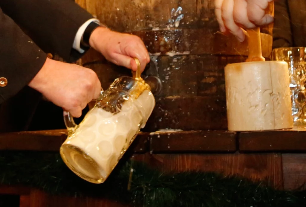 Anstich eines Bierfasses mit einem Holzschlegel, während frisches Bier schäumend in einen schräg gehaltenen Maßkrug gezapft wird.