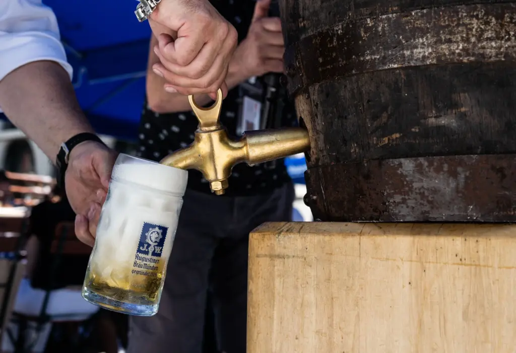 Nahaufnahme, wie eine Hand frisches Bier aus einem rustikalen Holzfass in einen Maßkrug mit Logo zapft. Der Krug ist bereits halbvoll mit hellem Bier und weißem Schaum.